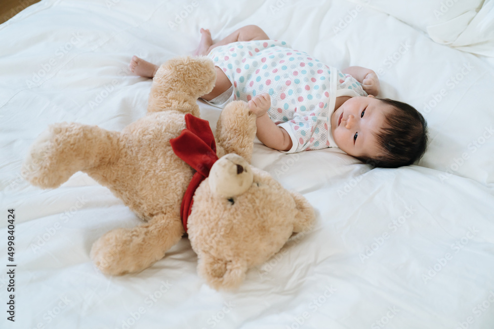 view of lovely asian baby lying on white bedding with teddy bear. female newborn in bodysuit is looking at stuffed toy innocently.