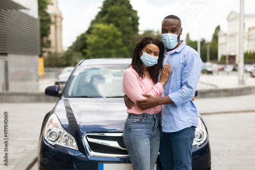 Happy black couple in masks standing near car and hugging