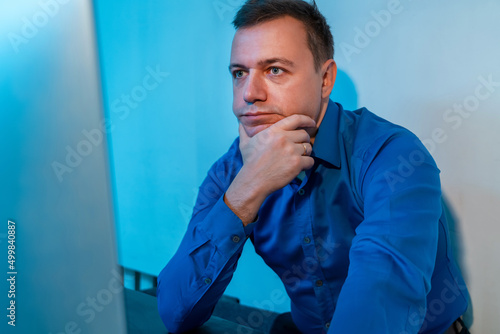 Millennial man sitting in office thinking about work project. Young businessman in blue shirt at startup business office space, working on desktop computer. Man working on computer at desk in home