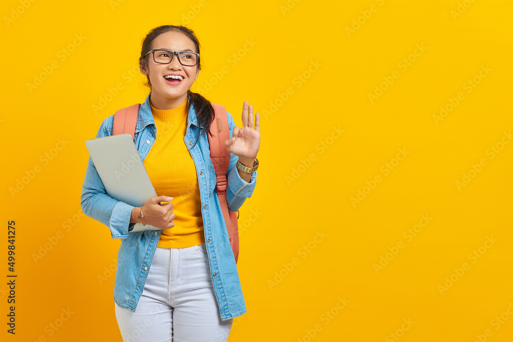 Portrait of cheerful young Asian woman student in casual clothes with backpack holding laptop and showing ok gesture with finger isolated on yellow background. Education in university college concept Portrait of cheerful young Asian woman student in casual clothes with backpack holding laptop and showing ok gesture with finger isolated on yellow background. Education in university college concept