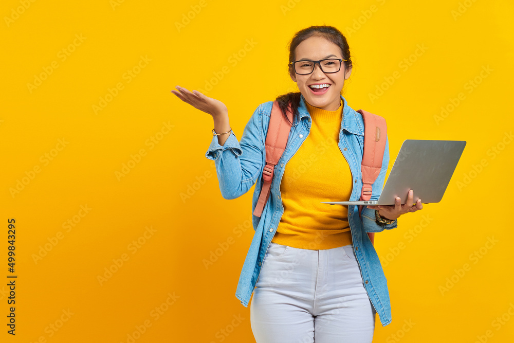 Portrait of smiling young Asian woman student in casual clothes with backpack showing copy space on palm and holding laptop isolated on yellow background. Education in university college concept Portrait of smiling young Asian woman student in casual clothes with backpack showing copy space on palm and holding laptop isolated on yellow background. Education in university college concept