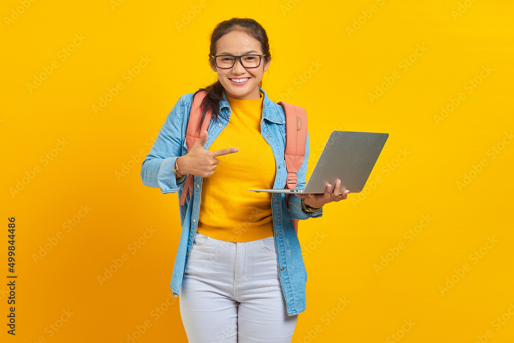 Portrait of smiling young Asian woman student in casual wear with backpack pointing at laptop with finger isolated on yellow background. Education in college university concept Portrait of smiling young Asian woman student in casual wear with backpack pointing at laptop with finger isolated on yellow background. Education in college university concept