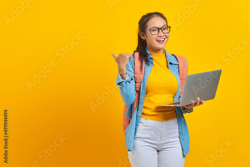 Portrait of smiling young Asian woman student in casual wear with backpack using laptop and pointing at copy space with finger isolated on yellow background. Education in college university concept
