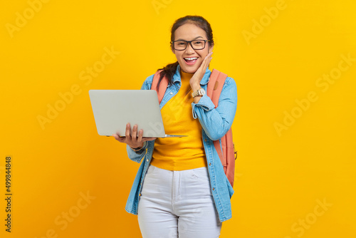 Portrait of excited young Asian woman student in casual clothes with backpack using laptop and touching cheek with hands isolated on yellow background. Education in college university concept