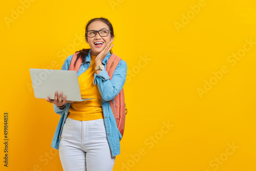 Portrait of excited young Asian woman student in casual clothes with backpack using laptop and touching cheek with hands isolated on yellow background. Education in college university concept