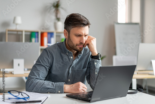 Serious middle aged businessman using laptop computer and reading reports, sitting at workplace in office photo
