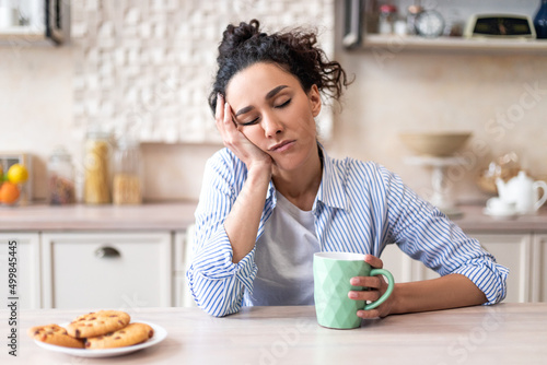 Sleepy young woman sitting at dining table in kitchen with closed eyes, holding cup while having breakfast photo