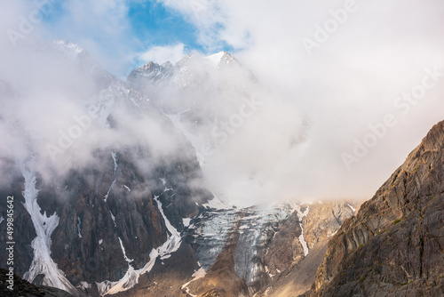 Scenic mountain landscape with snowy mountain top and glacier in dense low clouds in sunrise colors. Colorful mountain view to vertical glacier with icefall in morning sunlight in thick low clouds. © Daniil