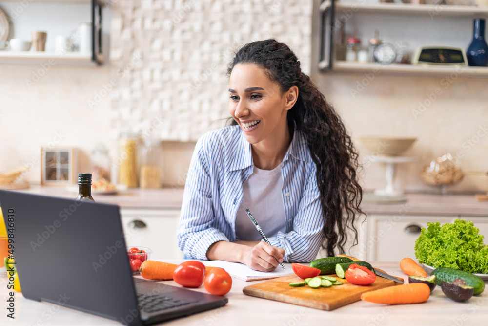 Writing down new recipe. Happy latin woman taking notes in copybook while watching cooking webinar on laptop