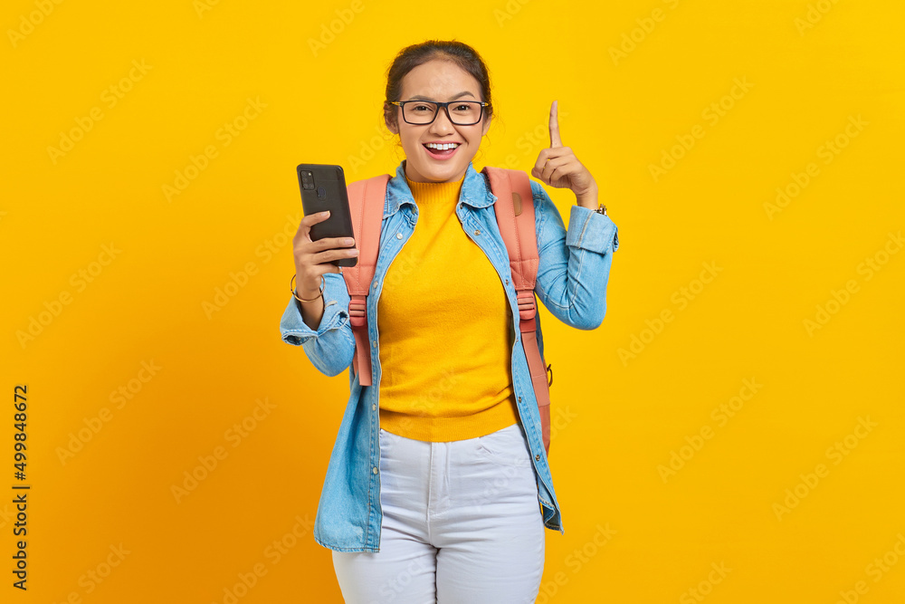Portrait of cheerful young Asian woman student in casual clothes with backpack holding mobile phone and pointing up space with fingers isolated on yellow background Portrait of cheerful young Asian woman student in casual clothes with backpack holding mobile phone and pointing up space with fingers isolated on yellow background