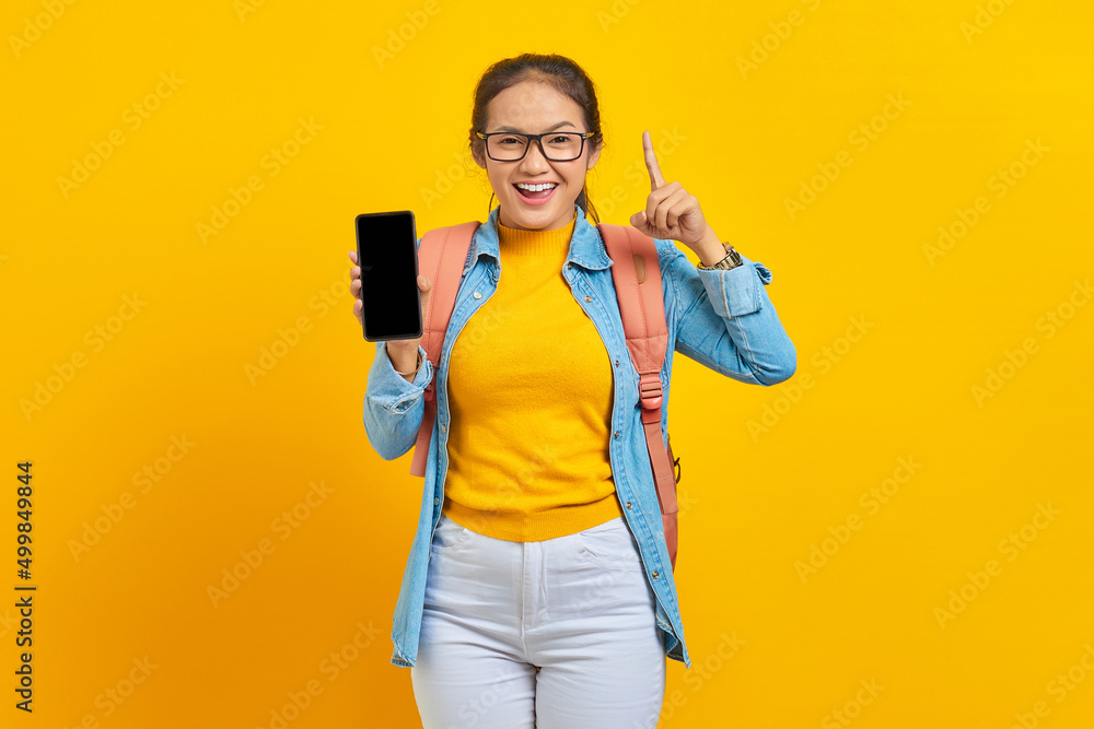Portrait of smiling young Asian woman student in casual clothes with backpack showing blank mobile phone screen and pointing fingers up isolated on yellow background Portrait of smiling young Asian woman student in casual clothes with backpack showing blank mobile phone screen and pointing fingers up isolated on yellow background