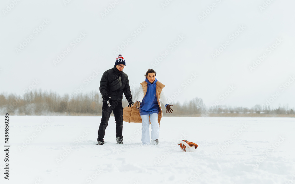 Positive man and woman with a smile on their faces having fun with a cute dog in the snow during a winter walk.