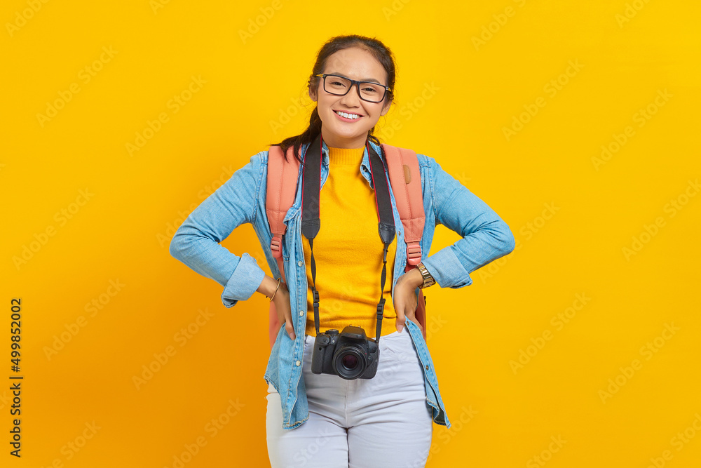 Portrait of cheerful young traveler Asian woman with backpack and camera in denim clothes isolated on yellow background. Passenger traveling on weekends. Air flight journey concept Portrait of cheerful young traveler Asian woman with backpack and camera in denim clothes isolated on yellow background. Passenger traveling on weekends. Air flight journey concept