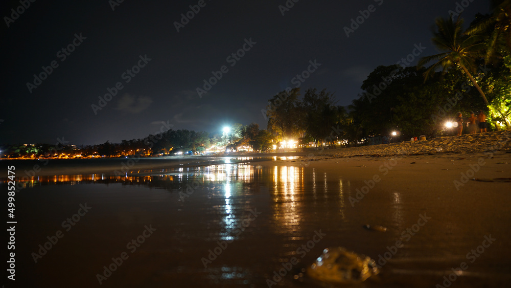 Night beach with a view of the clubs. Bright lights are burning reflecting in the sea. Fine wet sand. There is a transparent jellyfish. Palm trees grow. People are resting. Kata Beach, Phuket Night beach with a view of the clubs. Bright lights are burning reflecting in the sea. Fine wet sand. There is a transparent jellyfish. Palm trees grow. People are resting. Kata Beach, Phuket