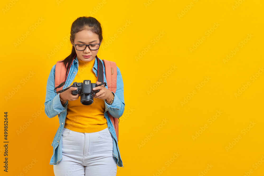 Portrait of pensive young Asian woman in denim clothes and looking at photo on camera isolated on yellow background. Passenger traveling on weekends. Air flight journey concept Portrait of pensive young Asian woman in denim clothes and looking at photo on camera isolated on yellow background. Passenger traveling on weekends. Air flight journey concept