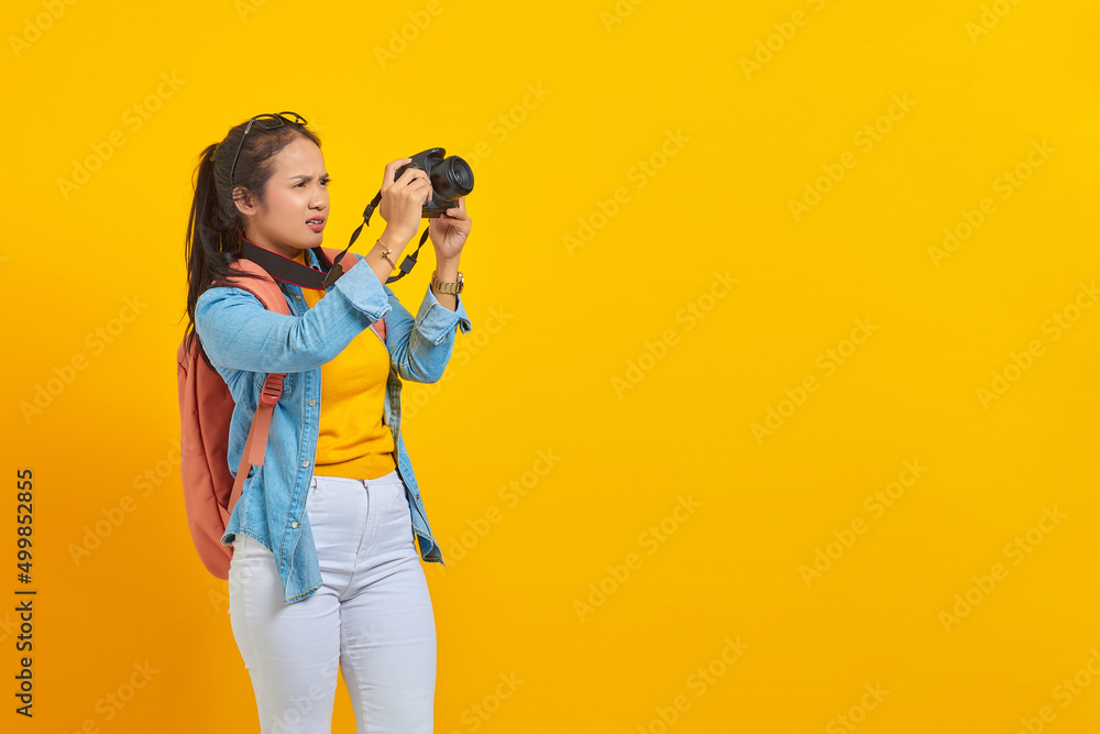 Obraz premium Portrait of surprised young Asian woman in denim clothes with backpack taking photo at camera profesional isolated on yellow background