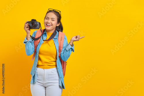 Portrait of smiling young Asian woman in denim clothes with backpack holding professional camera and pointing to copy space with finger isolated on yellow background