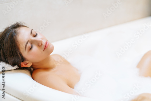 Young bare woman laying in white bubble bath with closed eyes side view.