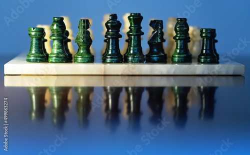 Dark green and ivory stone chess pieces on the chessboard and their reflection on the table. Blue background. 