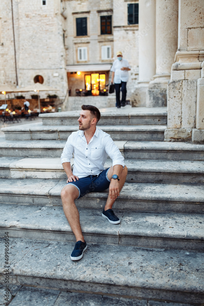 Handsome sexy man in white shirt sitting on the stairs in the old European city. Summer in Croatia. Dubrovnik city Handsome sexy man in white shirt sitting on the stairs in the old European city. Summer in Croatia. Dubrovnik city