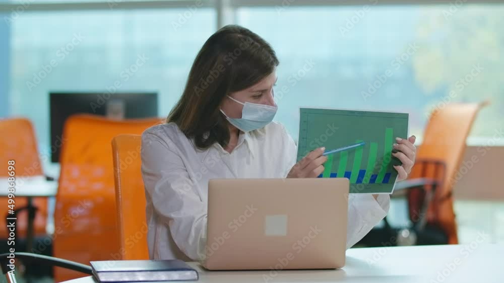 Serious intelligent woman in coronavirus face mask sitting at table with laptop showing profitability graph at web chat. Portrait of Caucasian manager conferencing online on pandemic lockdown Serious intelligent woman in coronavirus face mask sitting at table with laptop showing profitability graph at web chat. Portrait of Caucasian manager conferencing online on pandemic lockdown