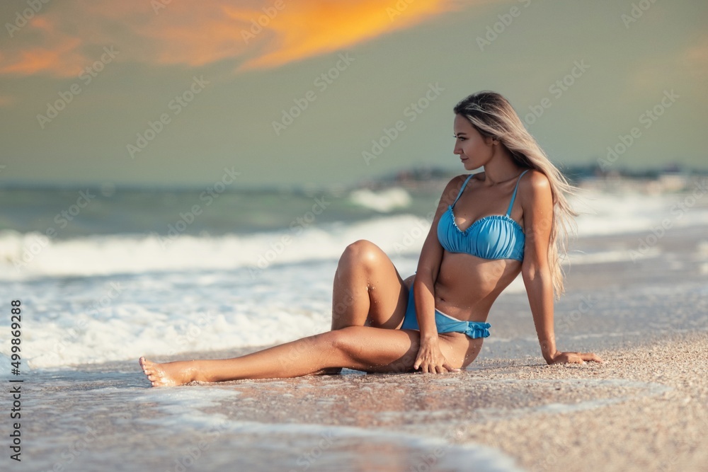 A girl with blond hair in a delicate swimsuit sits by the sea, enjoying the splash of waves A girl with blond hair in a delicate swimsuit sits by the sea, enjoying the splash of waves