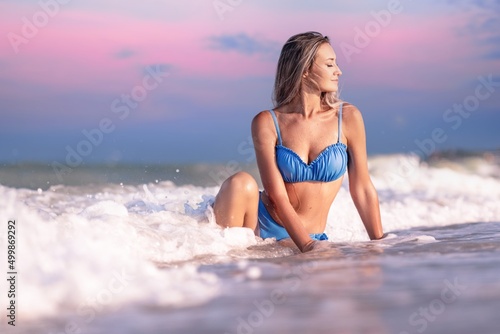A girl with blond hair in a delicate swimsuit sits by the sea  enjoying the splash of waves