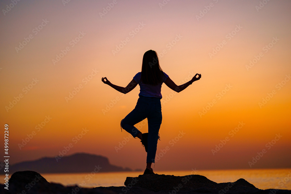 Back view of free calm bliss satisfied woman standing on top rock with yoga position against of idyllic sunset sky by the sea in a happy beautiful inspired moment of her life