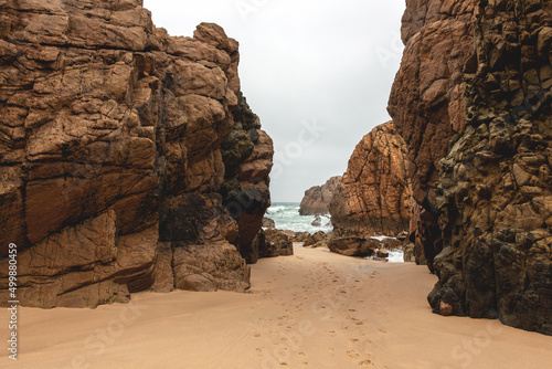 Landscape of Ursa beach through brown high rocks and stones with footprints in the sand towards turquoise water with waves of the coast of the atlantic ocean in cloudy weather, Portugal