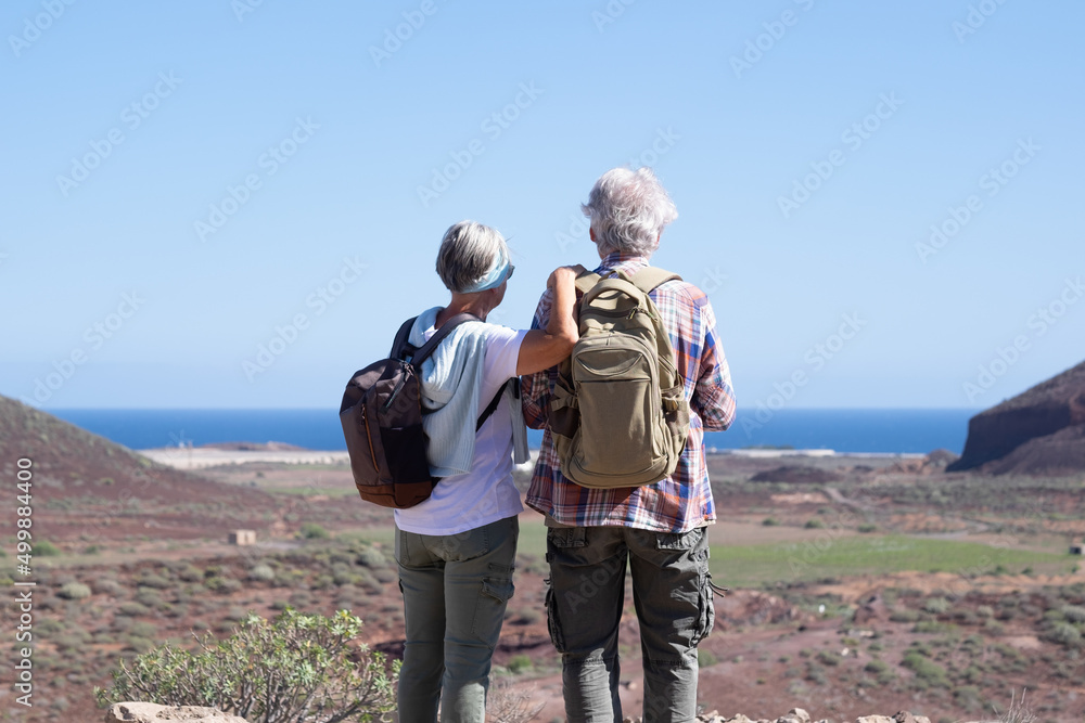 Active senior couple with backpacks in outdoors excursion hiking in mountain looking at horizon enjoying healthy lifestyle.  Scenic view of sea and mountain background