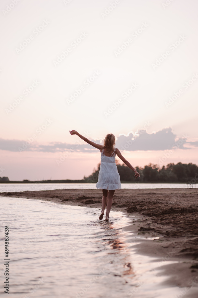 A young woman walks along the beach. Photo back view of a beautiful slender happy blonde in a white summer dress on the river bank against the background of sunset and clouds. 