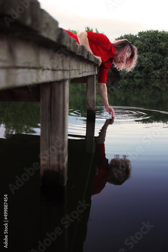 A young woman in a red dress leans over the bridge and touches the surface of the village lake with her hand. Outdoor recreation. Vacation in the village. Enjoying nature