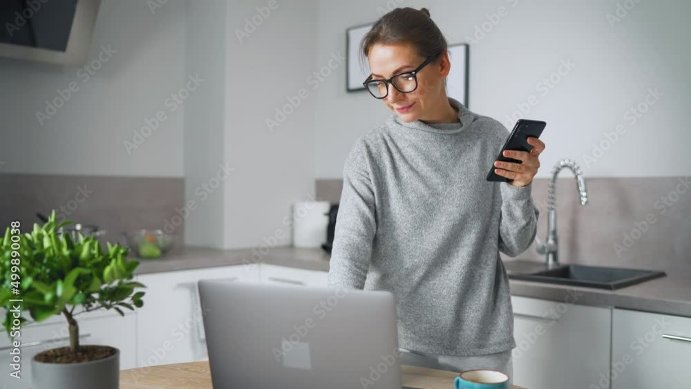 Woman with smartphone in hand standing in home cozy kitchen use laptop do freelance work, looking focused deep lost in thoughts. Concept of remote work. Home Office Woman with smartphone in hand standing in home cozy kitchen use laptop do freelance work, looking focused deep lost in thoughts. Concept of remote work. Home Office