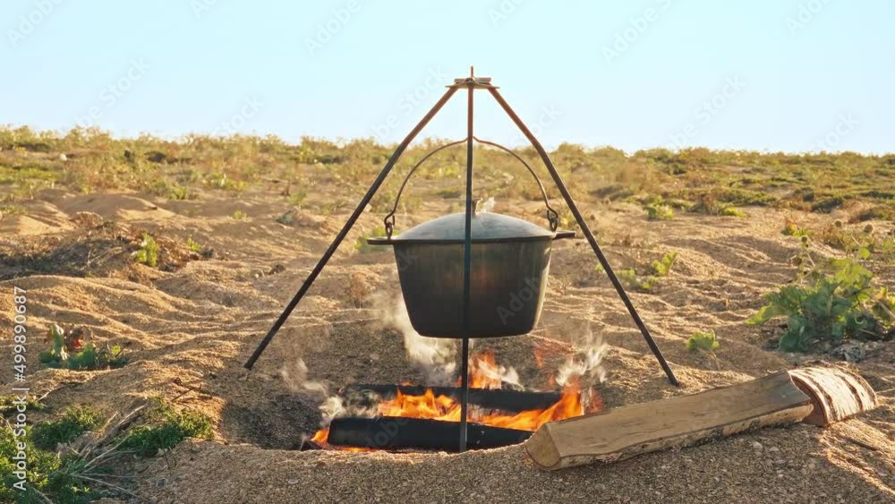 Cooking food on campfire. Tourist pot hangs over campfire at campsite on sandy beach. Concept of simple life by the sea under blue sky. Cooking food on campfire. Tourist pot hangs over campfire at campsite on sandy beach. Concept of simple life by the sea under blue sky.