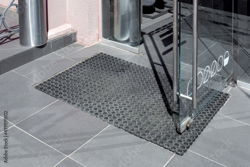 threshold made of gray ceramic stone tile at entrance to store with rubber foot mat and open glass door with metal handle and fastening loops at office building close up side view lit by sun, nobody.