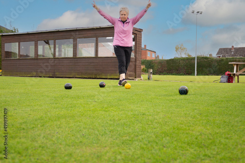 Happy female player signals that she has scored two points in crown green bowling competition by raising her arms, as she enjoys playing bowls outdoors on a warm Spring evening.