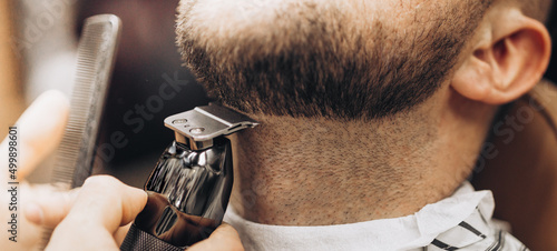 Close up cropped photo of a hairdresser`s work for a handsome young guy at the barber shop. He is doing styling with the electric shaver for his red beard photo