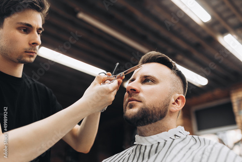 Fashion barber dressed in a black clothes tidies up men's beard and scissors it in the barbershop