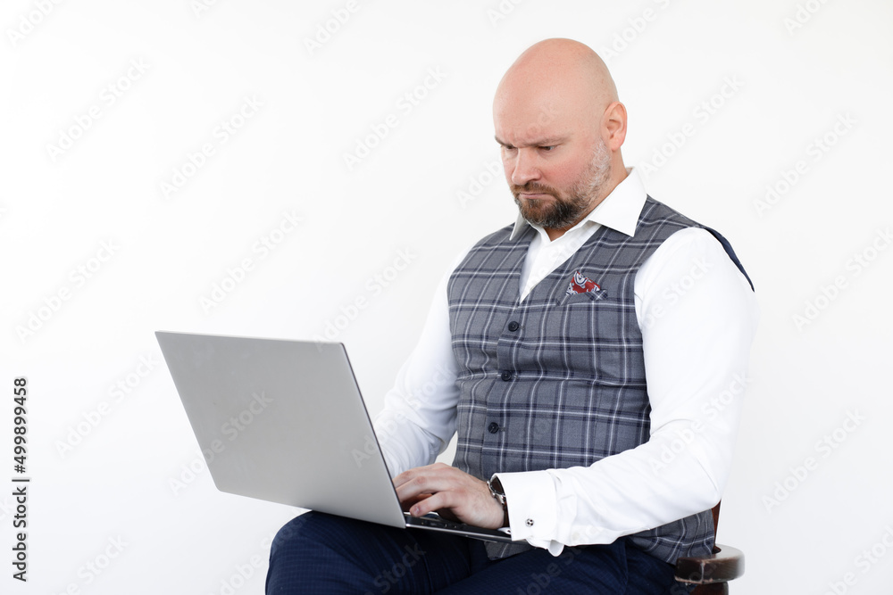 Portrait of serious bald businessman in grey vest, blue jeans, white shirt, sitting on chair, holding laptop,working. Portrait of serious bald businessman in grey vest, blue jeans, white shirt, sitting on chair, holding laptop,working.