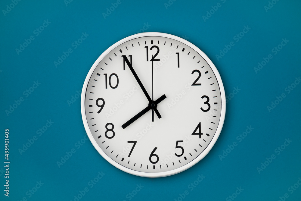 A white wall clock hangs on a blue wall with copy space and a central composition. A white wall clock hangs on a blue wall with copy space and a central composition.
