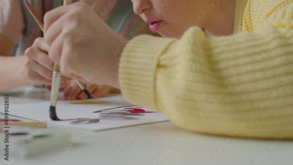 Close up tilt up shot of cheerful girl with Down syndrome painting picture on paper with assistance of female teacher and smiling during art lesson at home Close up tilt up shot of cheerful girl with Down syndrome painting picture on paper with assistance of female teacher and smiling during art lesson at home