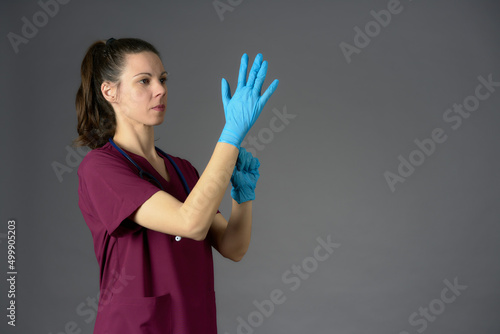 brunette nurse in purple uniform with stethoscope putting on latex medical gloves on gray background