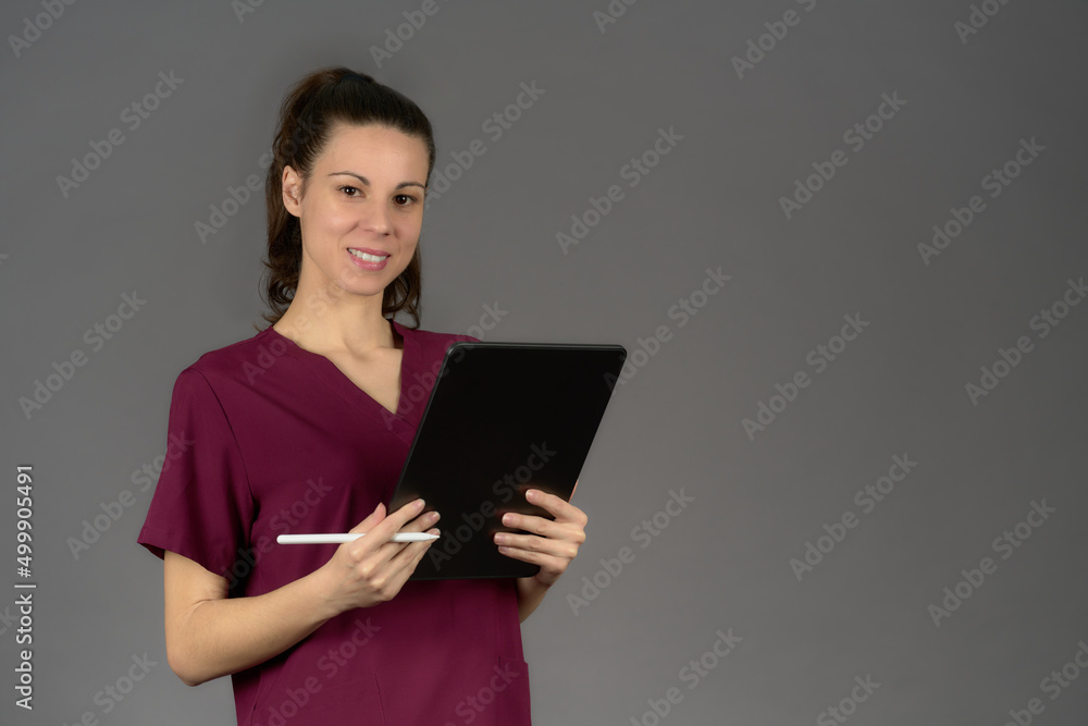 Fototapeta premium smiling female nurse in purple uniform looking at camera with digital tablet report on gray background