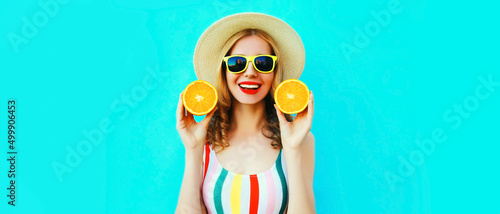 Summer portrait of happy smiling young woman with fresh slices of orange fruits wearing straw hat, sunglasses on blue background