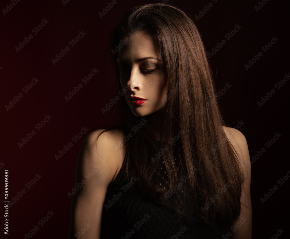 Beautiful bright makeup woman with red lipstick and long brown healthy hair with half face in shadow looking down on dark red shadow background. Closeup