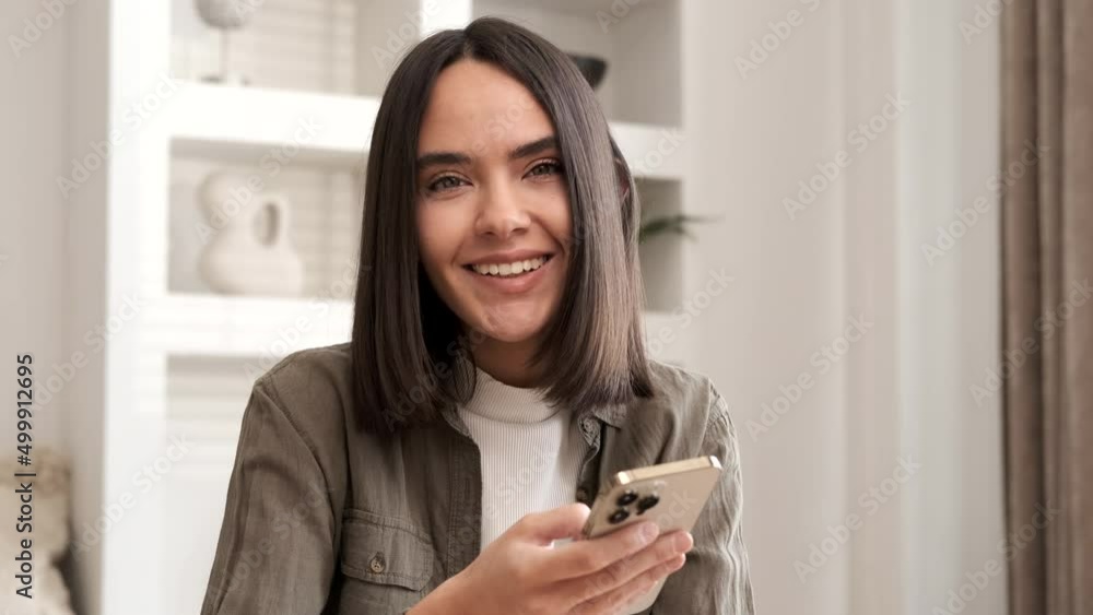 Happy woman smiling looking at camera. Home office interior. Business woman closeup portrait. Girl using smartphone internet surfing communication online. Pretty woman holding phone. Slow motion Happy woman smiling looking at camera. Home office interior. Business woman closeup portrait. Girl using smartphone internet surfing communication online. Pretty woman holding phone. Slow motion
