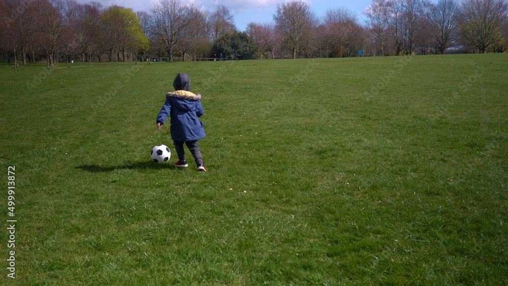 Happy Family Of Children Having Fun In Spring Park. Little Kid Run. Child Boy Dribbles Black White Classic Soccer Ball On Green Grass. People Playing Football. Childhood, Sport, Championship Concept Happy Family Of Children Having Fun In Spring Park. Little Kid Run. Child Boy Dribbles Black White Classic Soccer Ball On Green Grass. People Playing Football. Childhood, Sport, Championship Concept