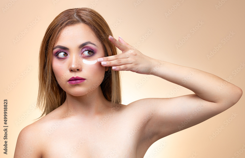Naklejka premium Close up portrait of a beautiful half naked woman with perfect, natural skin with fashion makeup. Applying cosmetic skincare cream, hydrated, lotion or mask on her face. Isolated over brown background