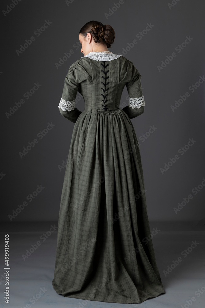 A Victorian working class woman wearing a checked bodice and skirt and standing alone against a studio backdrop A Victorian working class woman wearing a checked bodice and skirt and standing alone against a studio backdrop