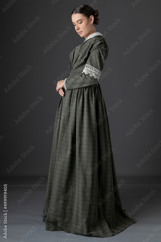 A Victorian working class woman wearing a checked bodice and skirt and standing alone against a studio backdrop A Victorian working class woman wearing a checked bodice and skirt and standing alone against a studio backdrop